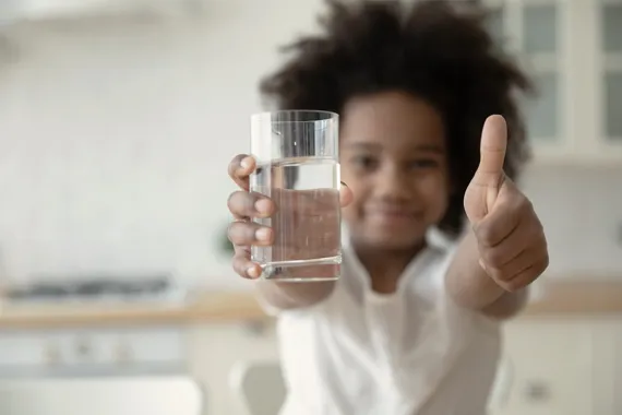 Flicka som håller i ett glas med vatten i ena handen och visar tummen upp med andra handen