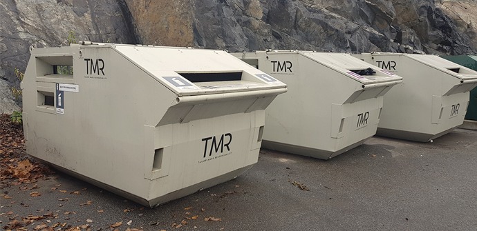 Tre gråa containrar för återvinning och insamling av avfall står på rad invid en bergvägg.