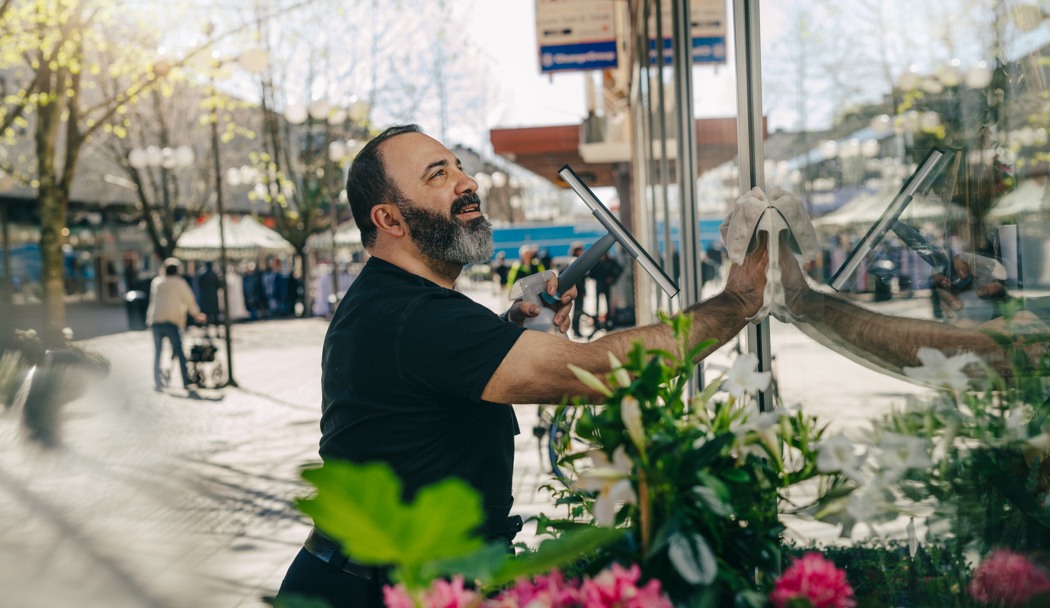 En man med skägg och svart t - shirt tvättar ett skyltfönster på en blomsteraffär. 