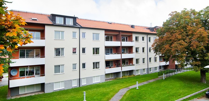Områdesbild på flerfamiljshus i tre våningar med gräsmatta och träd med gröna och gula löv utanför huset
